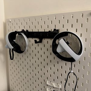 May include: White and black VR headset controllers mounted on a white pegboard with a black bracket. The controllers are suspended by their straps. The pegboard is attached to a white wall.