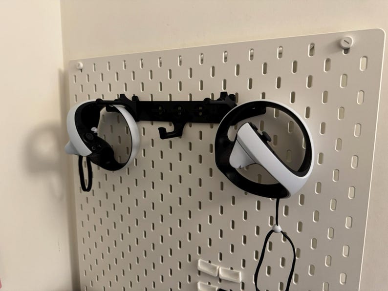 May include: White and black VR headset controllers mounted on a white pegboard with a black bracket. The controllers are suspended by their straps. The pegboard is attached to a white wall.