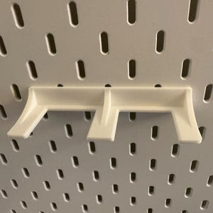 May include: A white pegboard shelf bracket, designed to fit into the slotted holes of a pegboard. The bracket has a simple, functional design with two angled supports and a horizontal base.