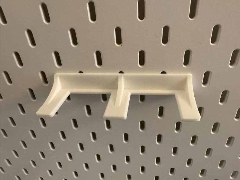 May include: A white pegboard shelf bracket, designed to fit into the slotted holes of a pegboard. The bracket has a simple, functional design with two angled supports and a horizontal base.