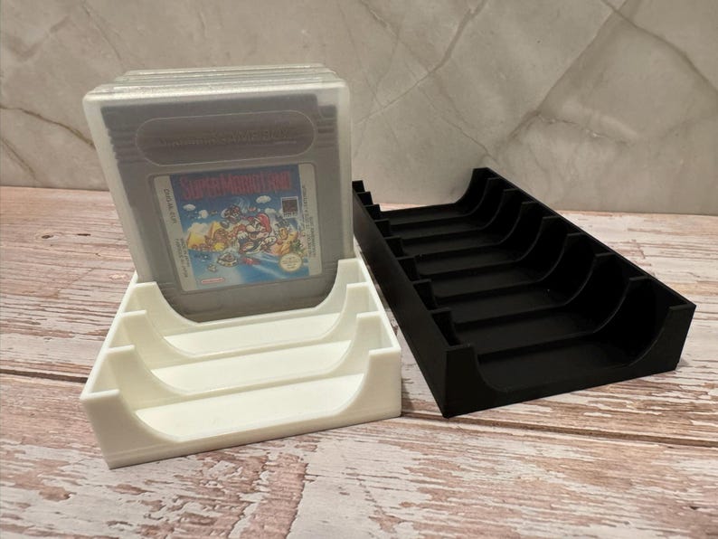 K&ouml;nnte beinhalten: Zwei 3D-gedruckte Spielkassettenhalter, einer wei&szlig; und einer schwarz. Eine Super Mario Land Game Boy-Kassette befindet sich im wei&szlig;en Halter. Der schwarze Halter ist leer. Diese Halter sind zum Aufbewahren und Pr&auml;sentieren von Retro-Videospielkassetten konzipiert.