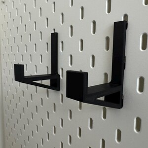 Op de afbeelding: Twee zwarte, L-vormige beugels voor een pegboard. De beugels zijn gemaakt van een solide materiaal en hebben een eenvoudig, functioneel ontwerp. Ze zijn gemonteerd op een wit pegboard met gelijkmatig verdeelde gaten.