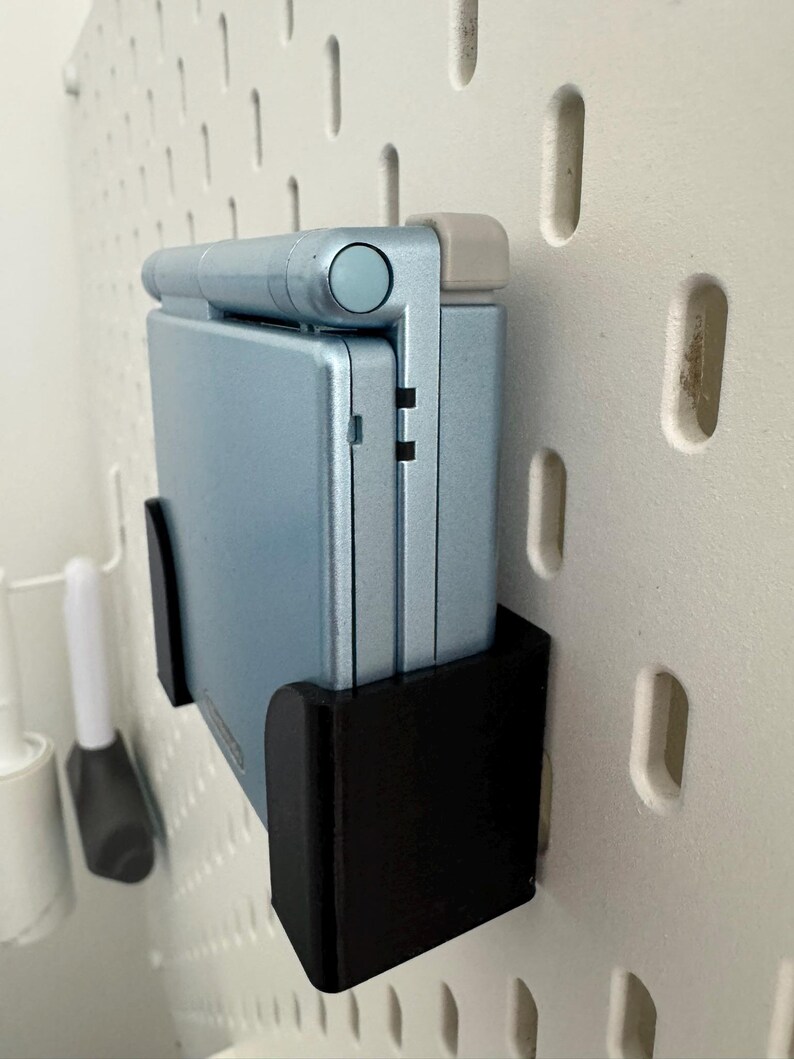 Pu&ograve; includere: Una console per videogiochi portatile Game Boy Advance SP di colore azzurro chiaro &egrave; montata su un supporto da parete nero. La console &egrave; chiusa e lo schermo &egrave; rivolto verso lo spettatore.