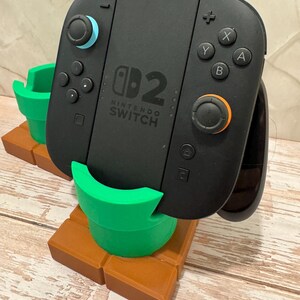 May include: Black Nintendo Switch controller with blue and orange accents, on a brown and green stand. The stand is designed to look like a Mario pipe and brick platform. The Nintendo Switch logo is visible on the controller.