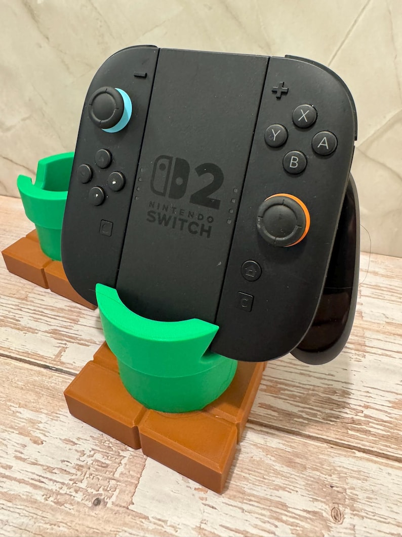 May include: Black Nintendo Switch controller with blue and orange accents, on a brown and green stand. The stand is designed to look like a Mario pipe and brick platform. The Nintendo Switch logo is visible on the controller.