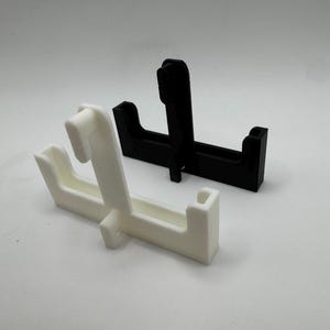 Op de afbeelding: Twee 3D-geprinte displaystandaards. E&eacute;n is wit, de andere is zwart. Elke standaard heeft een rechthoekige basis met een verticale steun en een gebogen bovenkant. De standaards zijn ontworpen om items te tonen.