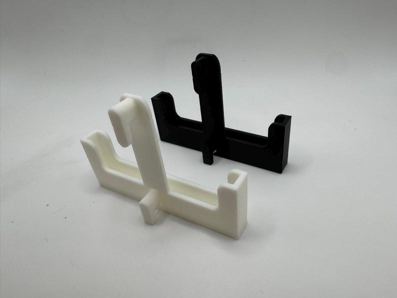 Op de afbeelding: Twee 3D-geprinte displaystandaards. E&eacute;n is wit, de andere is zwart. Elke standaard heeft een rechthoekige basis met een verticale steun en een gebogen bovenkant. De standaards zijn ontworpen om items te tonen.