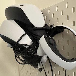 May include: A white and black VR headset and controller are mounted on a white pegboard. The headset has a curved design with a black top and white front. The controller is white with black buttons and a circular design.