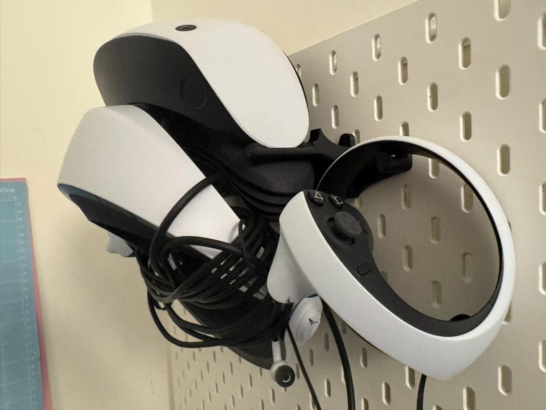 May include: A white and black VR headset and controller are mounted on a white pegboard. The headset has a curved design with a black top and white front. The controller is white with black buttons and a circular design.