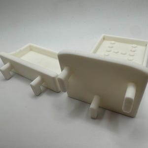 May include: Two off-white ceramic trays with rectangular bases and raised edges. Each tray has four short, rectangular legs. One tray has a series of small, rounded indentations on its surface.