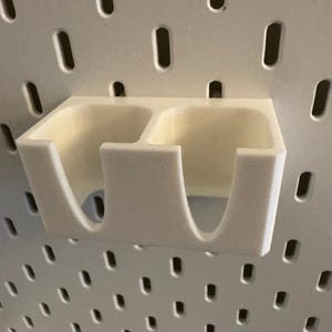 Puede incluir: Un organizador de panel perforado de color crema, impreso en 3D, con dos compartimentos. El organizador est&aacute; montado en un panel perforado blanco con agujeros espaciados. El dise&ntilde;o presenta bordes redondeados y una est&eacute;tica minimalista.