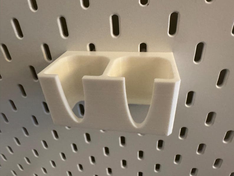 Puede incluir: Un organizador de panel perforado de color crema, impreso en 3D, con dos compartimentos. El organizador est&aacute; montado en un panel perforado blanco con agujeros espaciados. El dise&ntilde;o presenta bordes redondeados y una est&eacute;tica minimalista.