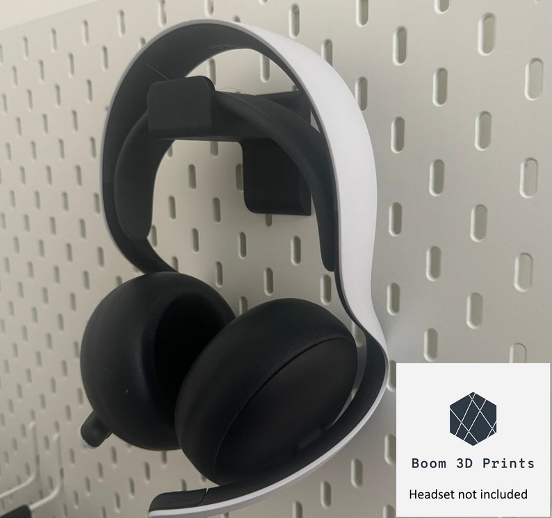 Pu&ograve; includere: Una cuffia nera e bianca appesa a un supporto a parete nero stampato in 3D. La cuffia &egrave; montata su una pannello forato bianco. L'immagine include il testo "Boom 3D Prints" e "Headset not included".