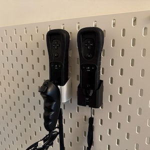 Puede incluir: Dos mandos de videojuegos negros están montados en una pared perforada blanca. Un mando está en un soporte blanco y el otro en un soporte negro. Los mandos tienen botones y almohadillas direccionales. También hay un mando negro con un joystick.