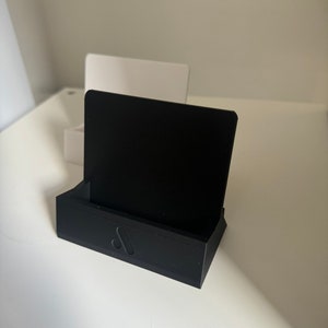 May include: A black business card holder with a rectangular base and a vertical back. The holder has a minimalist design with a small, stylized logo on the front. A black card is in the holder.
