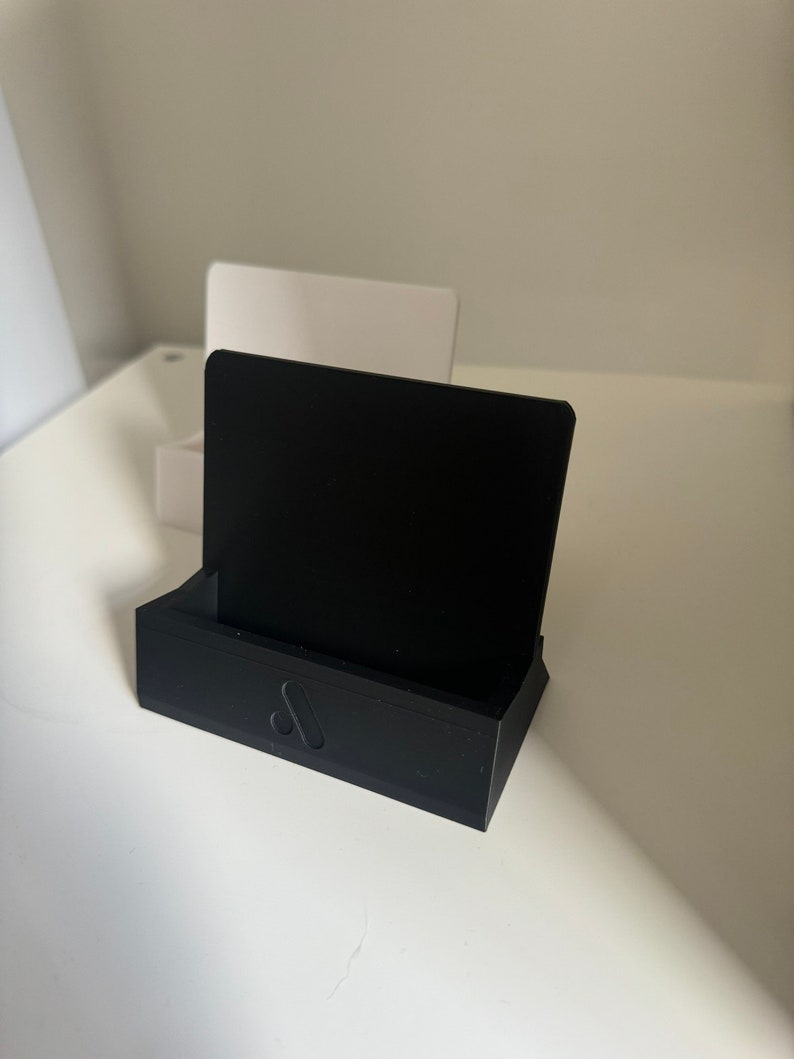 May include: A black business card holder with a rectangular base and a vertical back. The holder has a minimalist design with a small, stylized logo on the front. A black card is in the holder.