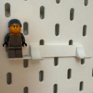 May include: A white plastic pegboard shelf with a blue and yellow Lego minifigure standing on it.