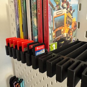 May include: Black 3D-printed shelf organizer holding Nintendo Switch game cartridges and game cases. The shelf is mounted on a white pegboard. Several game cartridges are visible, along with game cases for "Overcooked! 2" and "Wond Nintendo Switch 2 Edition".
