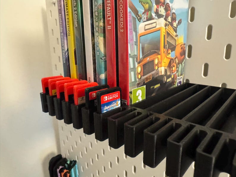 May include: Black 3D-printed shelf organizer holding Nintendo Switch game cartridges and game cases. The shelf is mounted on a white pegboard. Several game cartridges are visible, along with game cases for "Overcooked! 2" and "Wond Nintendo Switch 2 Edition".