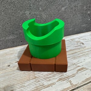 K&ouml;nnte beinhalten: Ein hellgr&uuml;nes Rohr, das an ein Videospiel erinnert, steht auf einer braunen, ziegelartigen Basis. Das Rohr hat eine gebogene &Ouml;ffnung und eine zylindrische Form. Die Basis ist quadratisch mit einem Ziegelmuster.