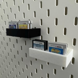 Op de afbeelding: Wit pegboard met twee planken met Nintendo 3DS-gamecartridges. De bovenste plank is zwart, de onderste wit. Spellen zijn onder meer "Detective Pikachu" en "Brain Training".