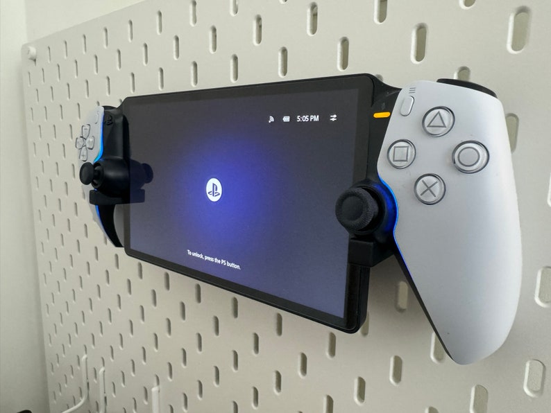 以下が含まれることがあります： 白いPlayStation 5 DualSenseコントローラーが、白いPlayStation Portal携帯型ゲーム機に取り付けられています。ゲーム機は、白いペグボードの壁に取り付けられています。ゲーム機の画面には、PlayStationのロゴと「ロックを解除するには、PSボタンを押してください。」というテキストが表示されています。