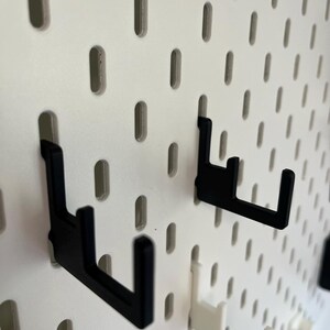 Puede incluir: Primer plano de un panel perforado blanco con varios ganchos negros y blancos. Los ganchos est&aacute;n dise&ntilde;ados para encajar en los agujeros del panel perforado, proporcionando una soluci&oacute;n de almacenamiento o exhibici&oacute;n. Los ganchos son de un material resistente.