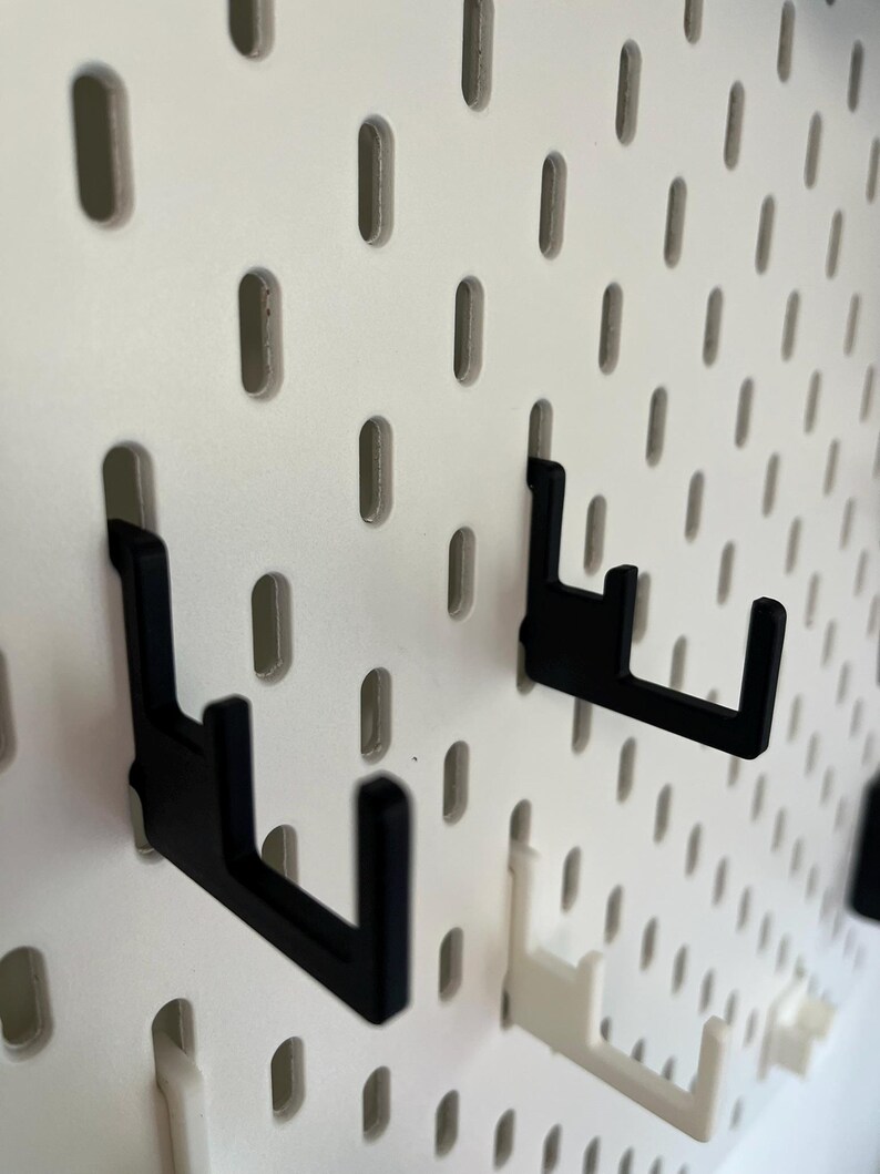 Puede incluir: Primer plano de un panel perforado blanco con varios ganchos negros y blancos. Los ganchos est&aacute;n dise&ntilde;ados para encajar en los agujeros del panel perforado, proporcionando una soluci&oacute;n de almacenamiento o exhibici&oacute;n. Los ganchos son de un material resistente.