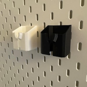 May include: Two 3D-printed pegboard organizers, one white and one black, are mounted on a white pegboard. The organizers have a rectangular shape with an angled top and a slot for holding items. The pegboard has evenly spaced holes.