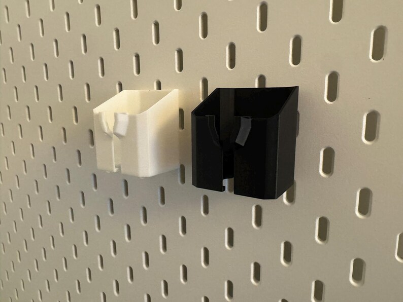 May include: Two 3D-printed pegboard organizers, one white and one black, are mounted on a white pegboard. The organizers have a rectangular shape with an angled top and a slot for holding items. The pegboard has evenly spaced holes.