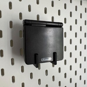 May include: A black Nintendo Game Boy Advance SP console mounted on a white pegboard. The console is held in place by a black bracket. The Nintendo logo is visible on the bracket.