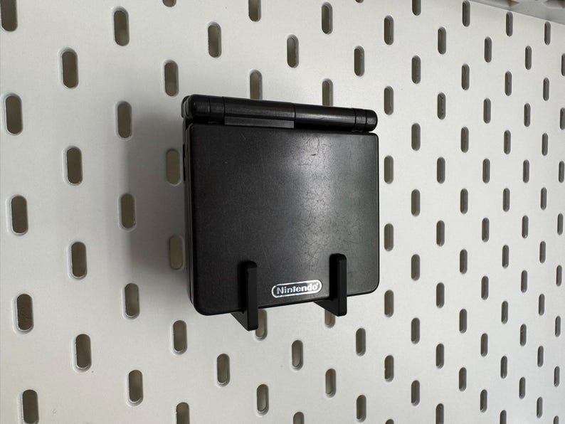 May include: A black Nintendo Game Boy Advance SP console mounted on a white pegboard. The console is held in place by a black bracket. The Nintendo logo is visible on the bracket.