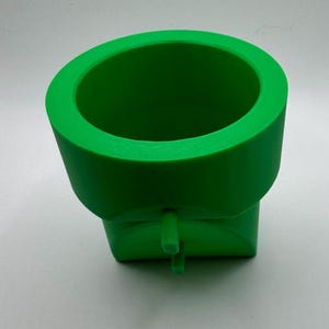 May include: Bright green plastic pipe fitting with a cylindrical top and a square base. The fitting has a small, cylindrical projection extending from the base. The object is likely used for plumbing or construction.