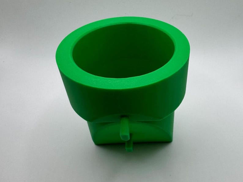 May include: Bright green plastic pipe fitting with a cylindrical top and a square base. The fitting has a small, cylindrical projection extending from the base. The object is likely used for plumbing or construction.