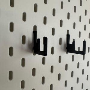 May include: Two black metal pegboard hooks are mounted on a white pegboard. The hooks are designed to hold items on the pegboard. The pegboard has evenly spaced oval holes.