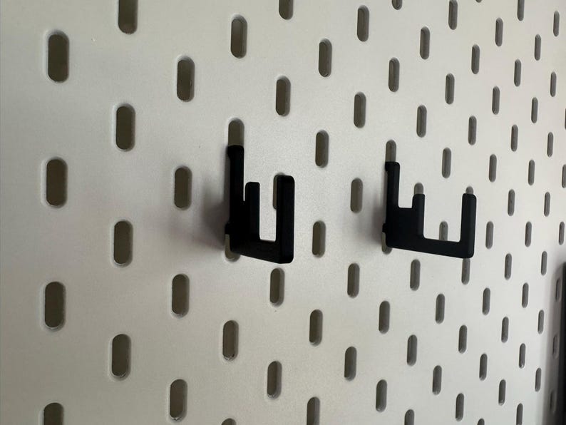 May include: Two black metal pegboard hooks are mounted on a white pegboard. The hooks are designed to hold items on the pegboard. The pegboard has evenly spaced oval holes.