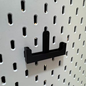 May include: Black pegboard hook accessory. The hook is designed to attach to a white pegboard with evenly spaced holes. The hook has a rectangular base with two upward-curving arms and a central vertical support.