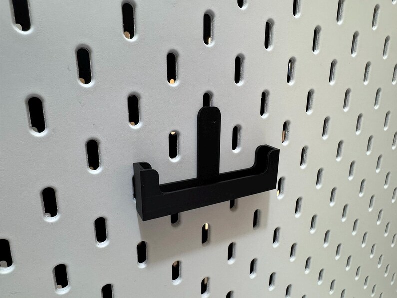 May include: Black pegboard hook accessory. The hook is designed to attach to a white pegboard with evenly spaced holes. The hook has a rectangular base with two upward-curving arms and a central vertical support.
