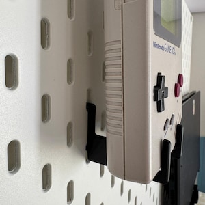 May include: A beige Nintendo Game Boy handheld console mounted on a white pegboard with black brackets. The console has a green screen, black directional pad, and pink buttons. The pegboard has a series of oval-shaped holes.