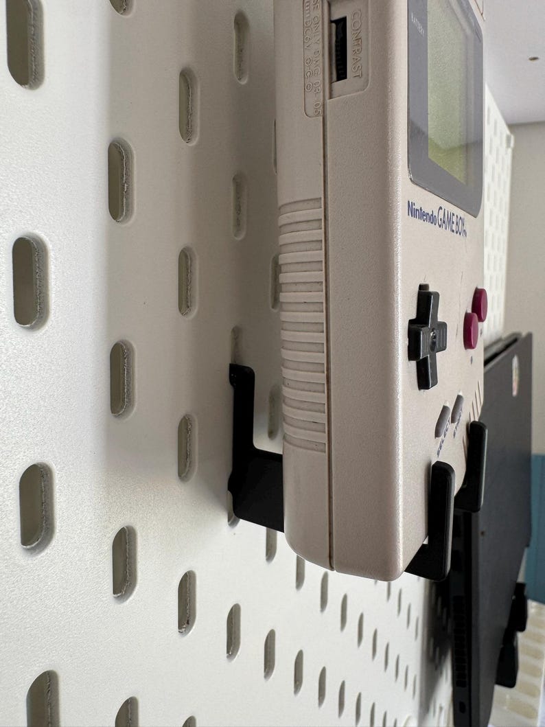 May include: A beige Nintendo Game Boy handheld console mounted on a white pegboard with black brackets. The console has a green screen, black directional pad, and pink buttons. The pegboard has a series of oval-shaped holes.