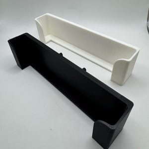 May include: Two rectangular storage containers, one black and one white, are displayed. Each container has a long, open top and raised sides. The black container is in the foreground, with the white container slightly behind it.