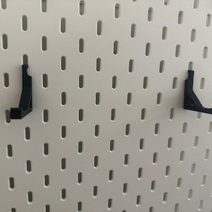 以下が含まれることがあります： 白いペグボードに2つの黒いプラスチック製の壁掛けフックが付いている
