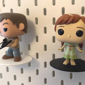 Op de afbeelding: Twee Funko Pop-figuren op displayschappen. De figuur links is een mannelijke figuur met bruin haar en een bruin en zwarte outfit. De figuur rechts is een vrouwelijke figuur met rood haar en een groene jurk.
