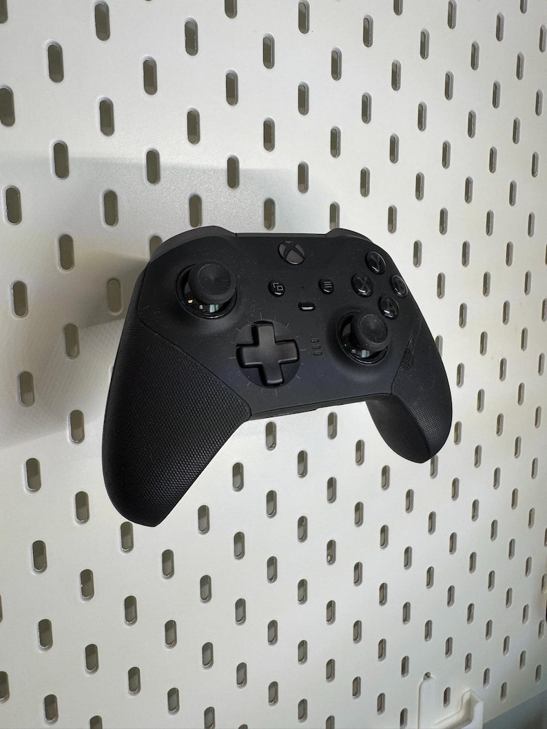 May include: A black Xbox controller with textured grips and several buttons. The controller includes a directional pad, two analogue sticks, and function buttons. It is mounted on a white pegboard.