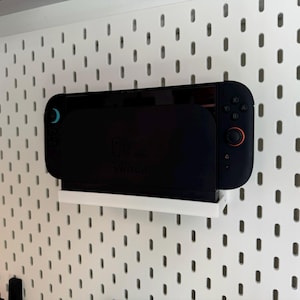 Puede incluir: Una consola Nintendo Switch negra est&aacute; montada en un estante blanco contra una pared de tablero perforado blanco. La consola tiene una pantalla negra y mandos con detalles en azul y naranja. El tablero perforado tiene un patr&oacute;n de agujeros espaciados.