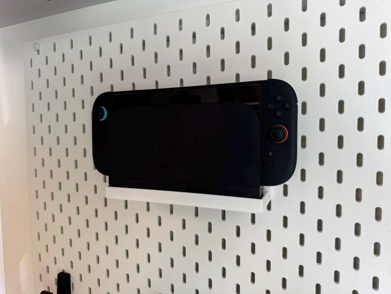 Puede incluir: Una consola Nintendo Switch negra est&aacute; montada en un estante blanco contra una pared de tablero perforado blanco. La consola tiene una pantalla negra y mandos con detalles en azul y naranja. El tablero perforado tiene un patr&oacute;n de agujeros espaciados.