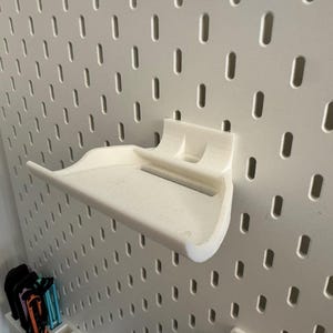 May include: A white 3D-printed shelf designed to attach to a pegboard. The shelf has a curved front and a flat surface for holding items. The pegboard is white with evenly spaced oval holes.