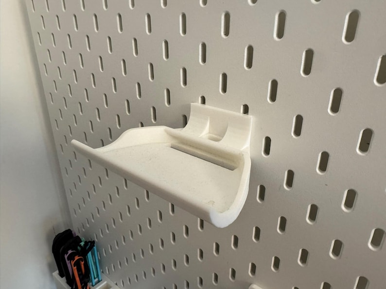 May include: A white 3D-printed shelf designed to attach to a pegboard. The shelf has a curved front and a flat surface for holding items. The pegboard is white with evenly spaced oval holes.