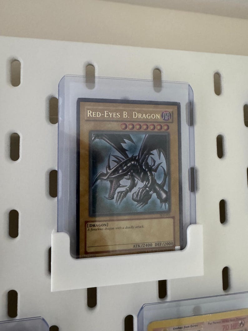 Pu&ograve; includere: Un protettore per carte in plastica trasparente con una carta da gioco di drago nero con gli occhi rossi al suo interno. La carta &egrave; intitolata "Red-Eyes B. Dragon" e ha uno sfondo nero con testo rosso e bianco. La carta &egrave; di tipo "Dragon" con un potere d'attacco di 2400 e un potere di difesa di 2000.