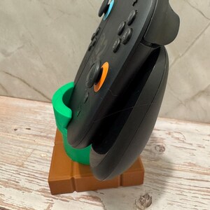 May include: Black video game controller in a green and brown stand. The stand is designed to resemble a green pipe on a brown brick base. The controller features blue and orange buttons.